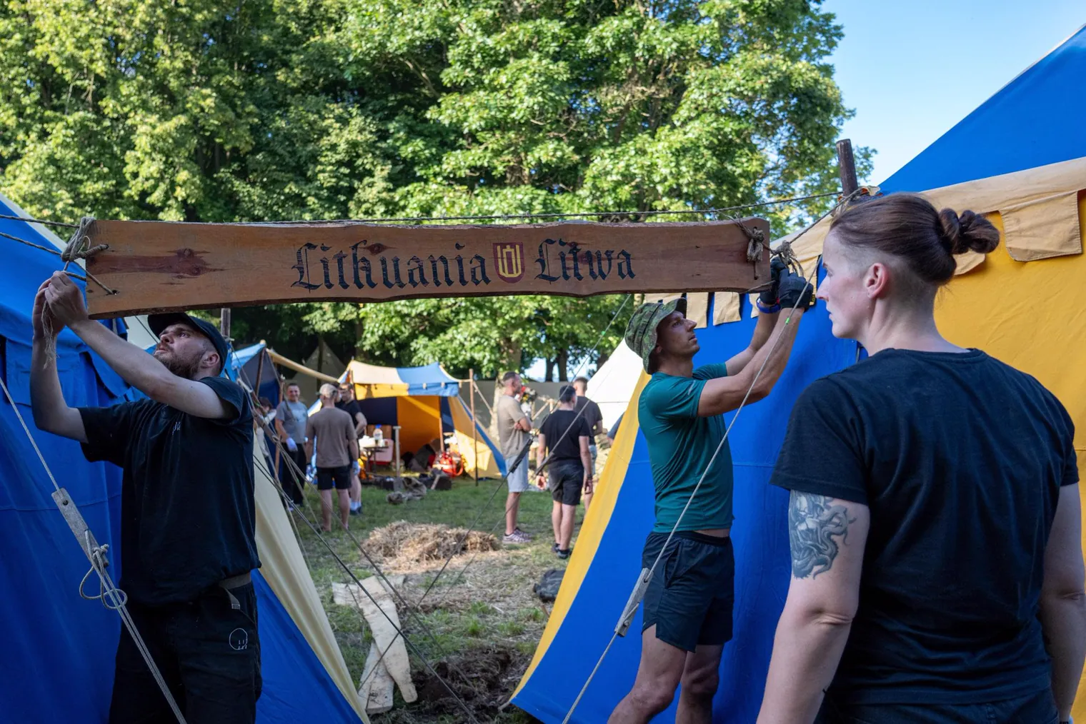 Tarp palapinių kabinama medinė lenta su užrašu „Lithuania · Litwa“ ir Gediminaičių stulpais